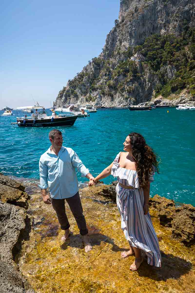 Couple holding hands and looking at each other while standing on yellow rocks with the beautiful Mediterranean sea as background