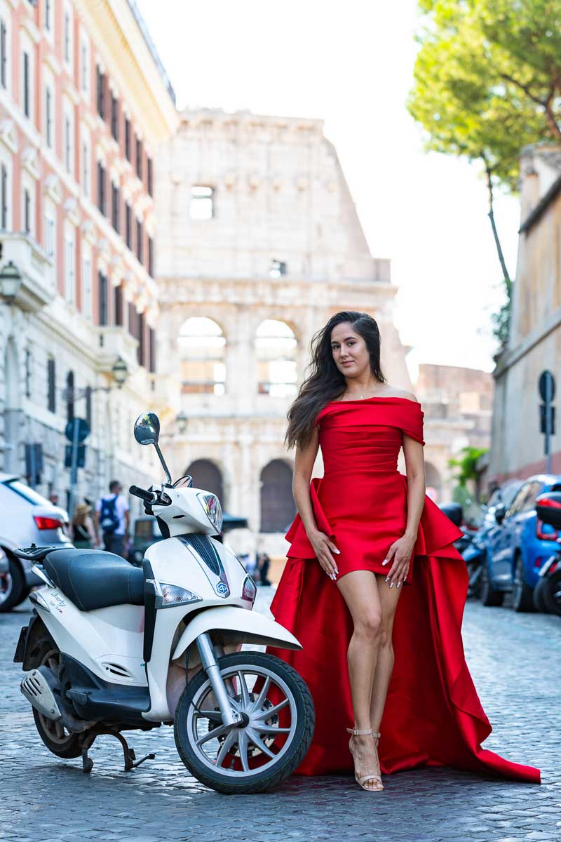Model wearing a bright red dress posing next to a scooter with the Coliseum background 