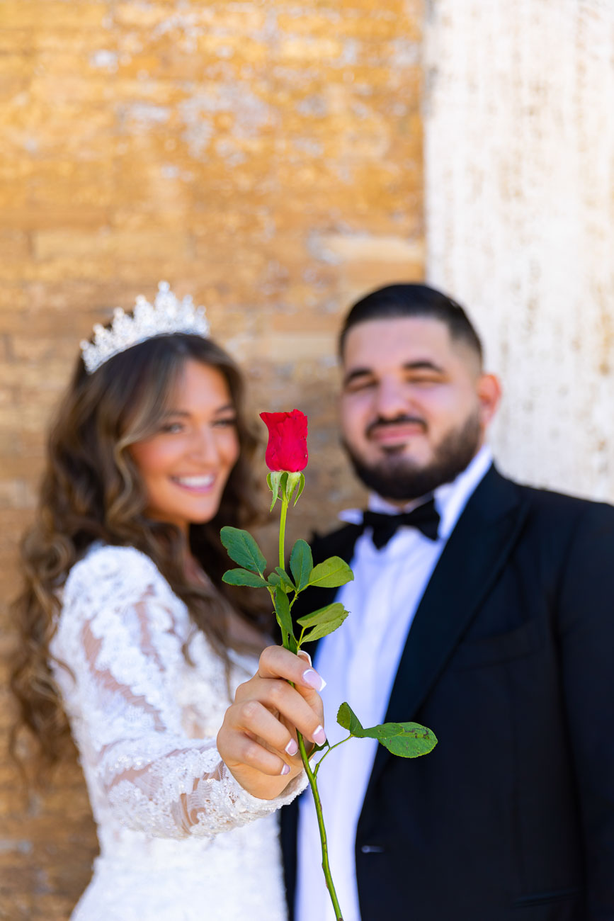 Bride holding a red rose in front for the camera with the bride and groom out of focus background