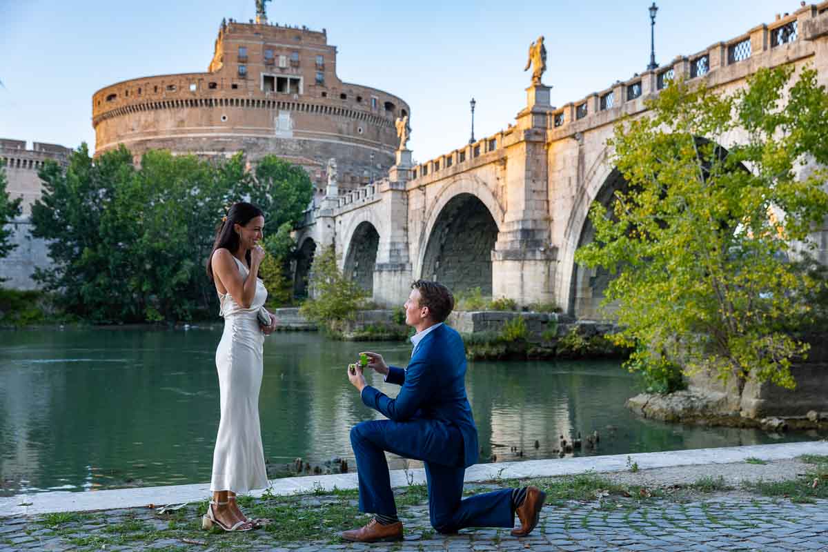 Kneeling down for a romantic surprise wedding proposal candidly photographed under the Castel Sant'Angelo bridge in Rome Italy