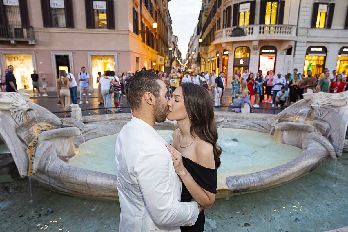 Kissing by the Barcaccia water fountain found at the bottom of the Spanish steps in Rome Italy as the lights start to turn on