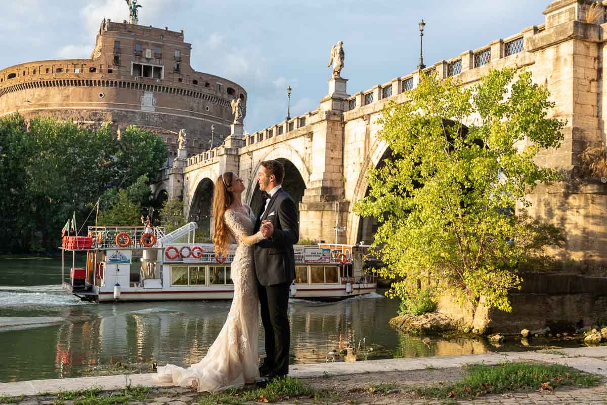 Groom and bride posed together under Castel Sant'Angelo bridge with a view Castle while a boat is rolling along river background