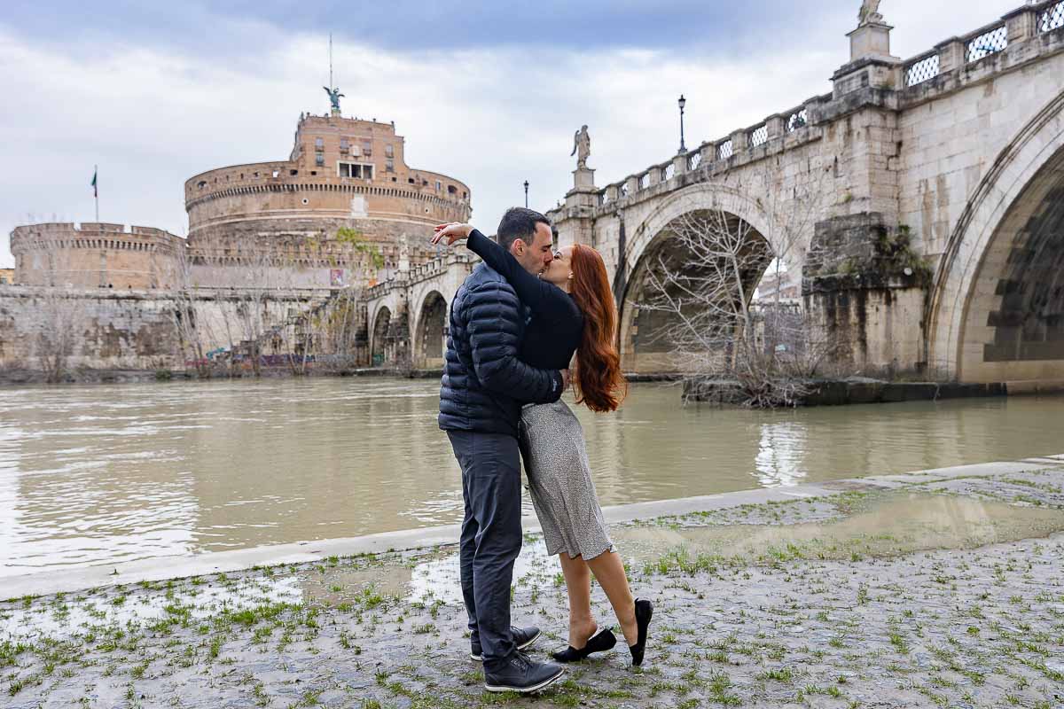 Engagement photo session down by the Tiber river level with the Castle and the Bridge in the far distance