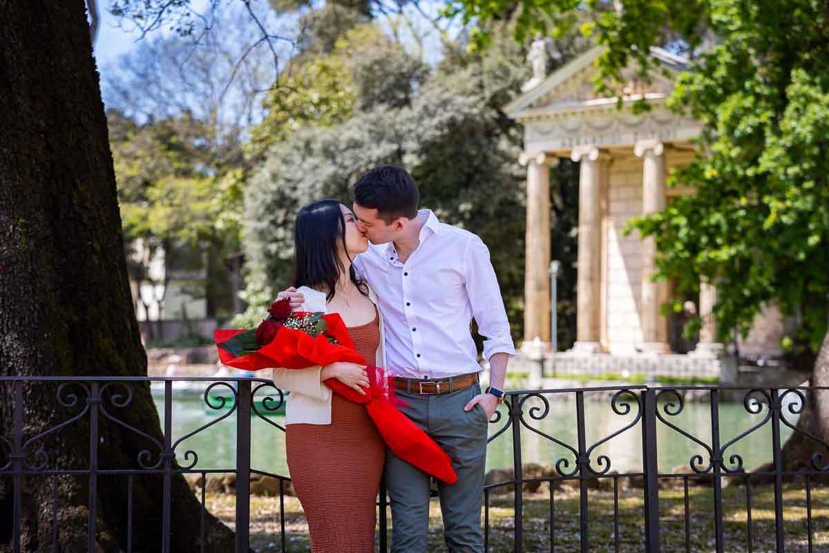 Kissing while holding a bouquet of red roses given to celebrate the engagement photos