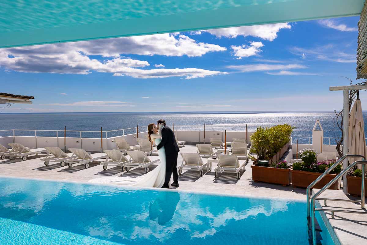 Bride and groom on a destination wedding before the amazing. And incredible view with white fluffy clouds and blue sky. Not to mention an amazing infinity swimming pool