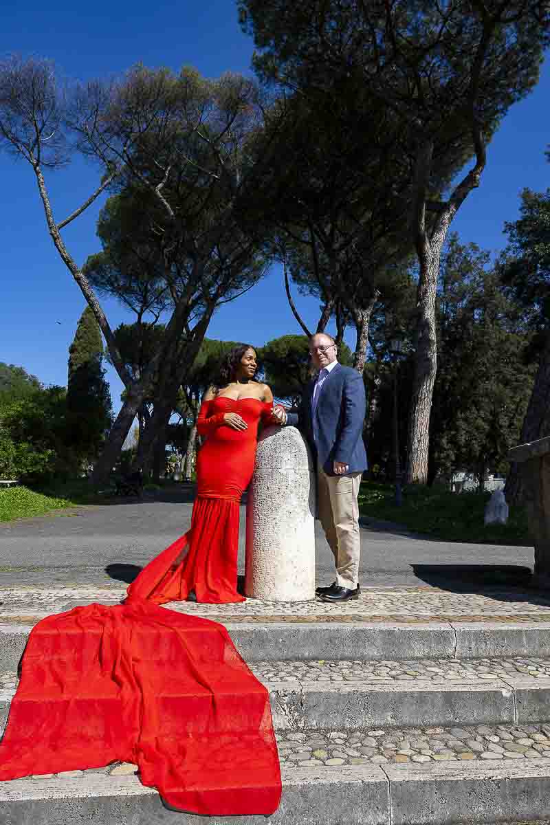 Posed couple long red dress tail falling over white marble steps park with Mediterranean pine trees photographer above by a lifestyle photographer session