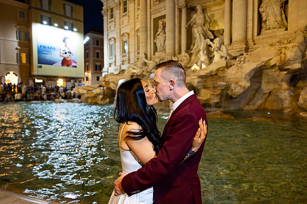 Kissing at night by Fontana di Trevi during a romantic photography session in wedding attire pictures taken right by the water redge
