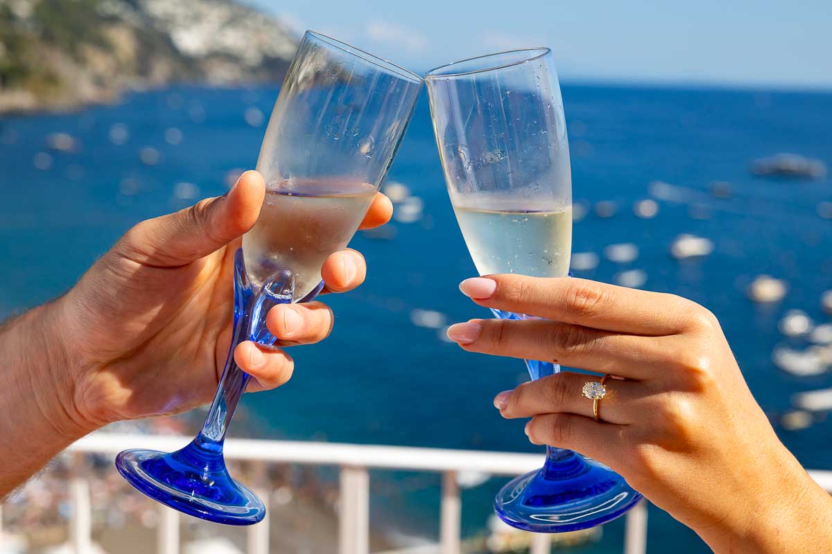 Engagement ring shown hand in hand while toasting with prosecco flute glasses tipping over the sea