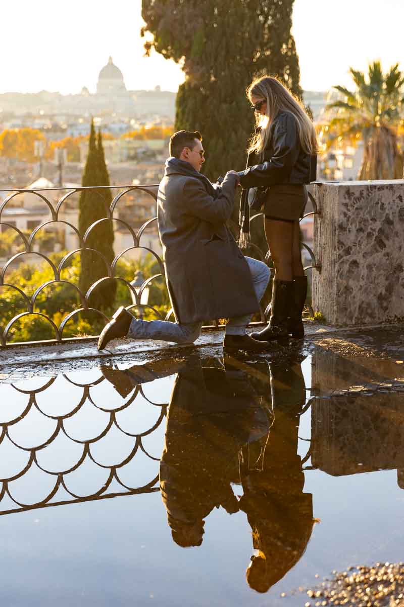 Proposing in Rome at the Pincio terrace panoramic outlook in front of a water puddle reflection and the typical metal fence 