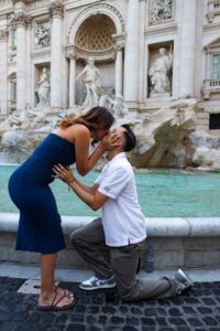 002-proposing-in-rome Newly engaged couple sharing a kiss while he remains on one knee beside the fountain, captured in a candid romantic photograph showing the joy of the surprise moment.