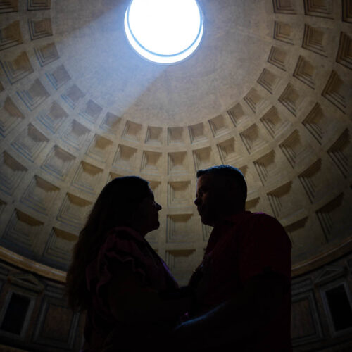 Silhouette of an engaged couple standing close beneath the dome with dramatic backlighting from the sunbeam above, captured in an artistic architectural photography composition