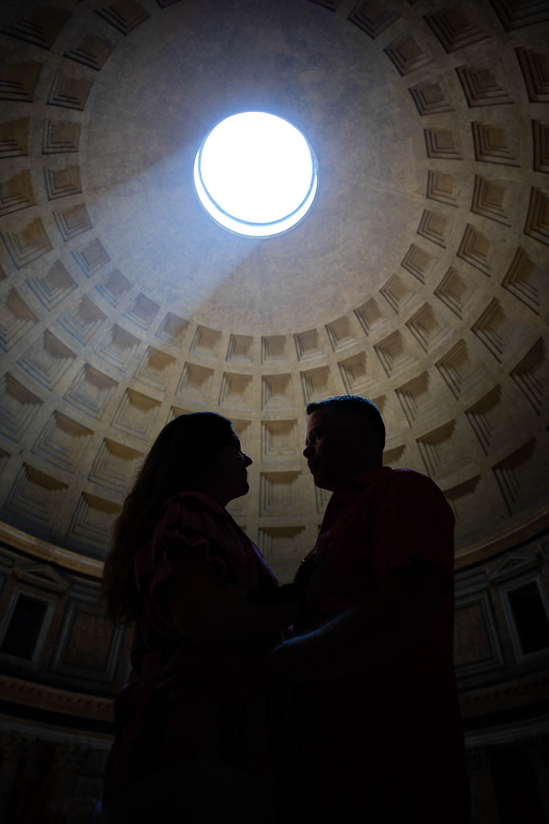 Silhouette of an engaged couple standing close beneath the dome with dramatic backlighting from the sunbeam above, captured in an artistic architectural photography composition