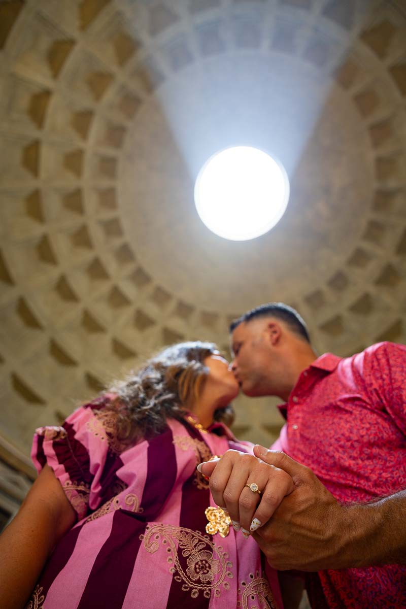 Newly engaged couple kissing while displaying the diamond ring beneath the dome opening with natural light creating a romantic architectural portrait