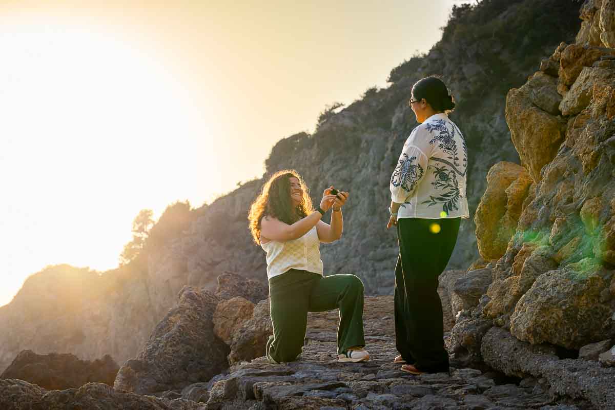 Backlit engagement photograph capturing the moment one woman kneels to ask her partner to marry her on the beach during the warm evening light.