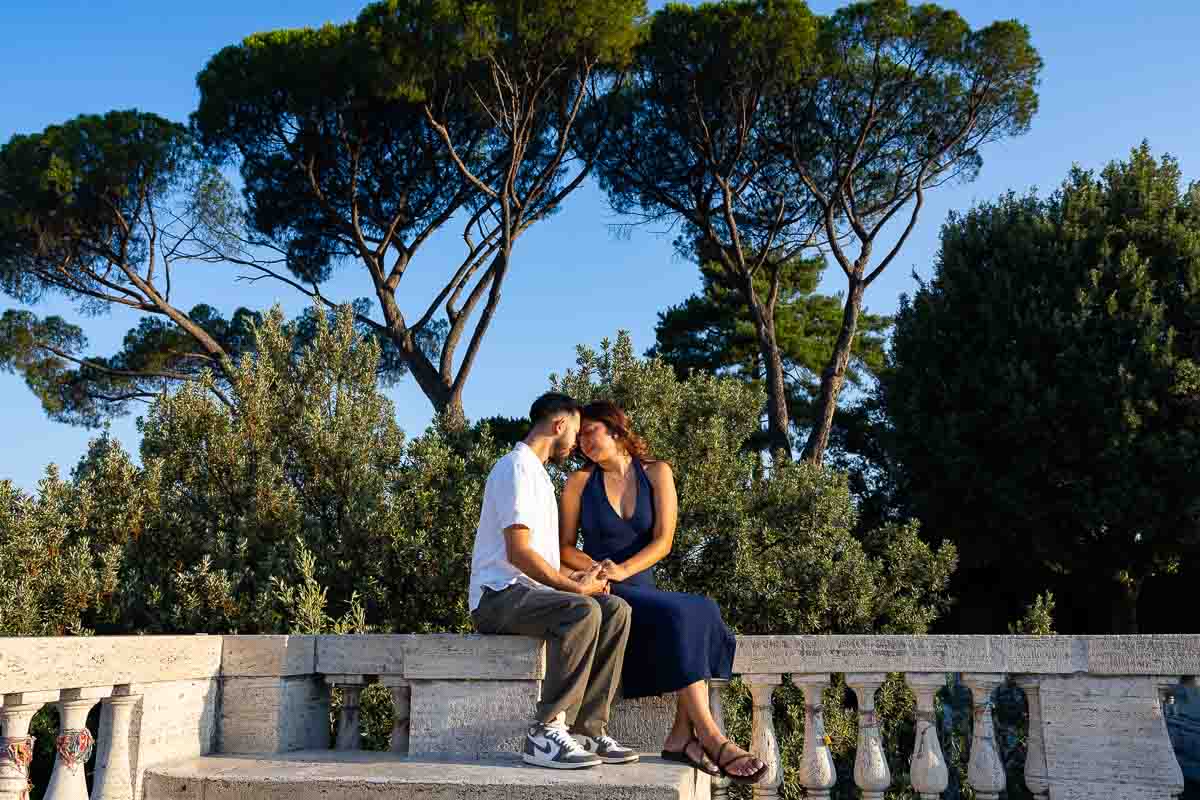 Couple seated facing each other with foreheads touching beneath Mediterranean pine trees, captured in an intimate romantic portrait with a natural engagement photoshoot style and soft outdoor atmosphere.