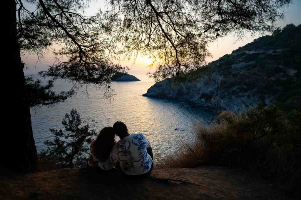 Panoramic sunset portrait of two women sitting side by side overlooking Cala del Gesso, celebrating their engagement with sweeping views across the Monte Argentario coastline.