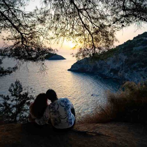 Panoramic sunset portrait of two women sitting side by side overlooking Cala del Gesso, celebrating their engagement with sweeping views across the Monte Argentario coastline.