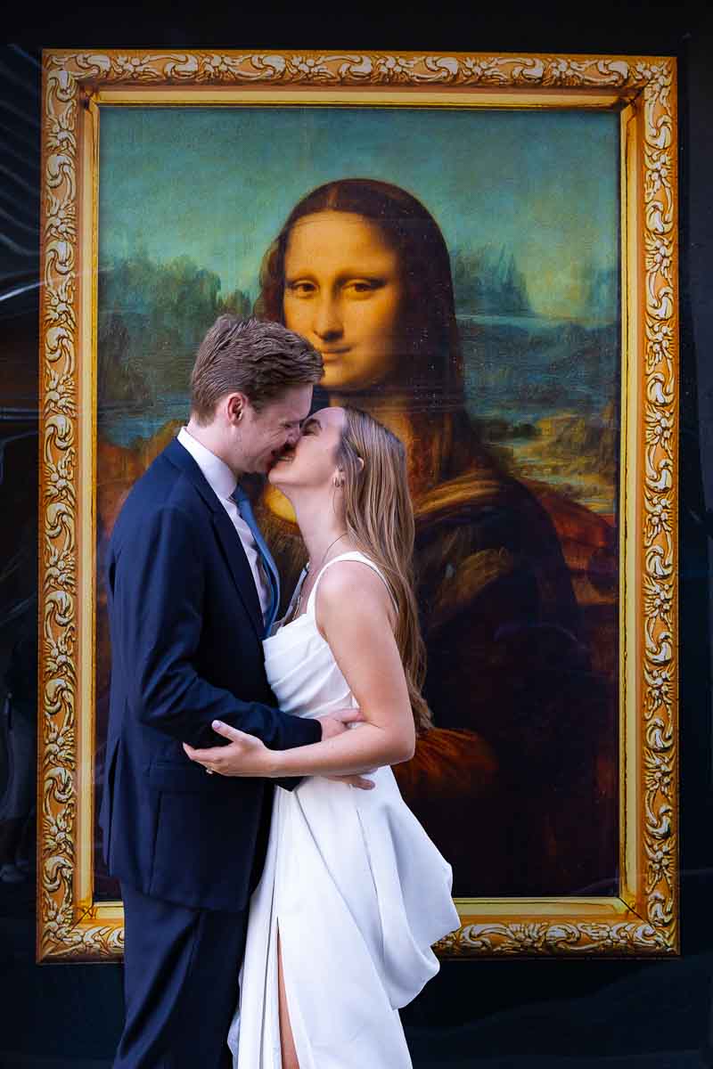 Bride and groom posing beside a replica of Leonardo da Vinci’s Mona Lisa, creating an artistic portrait with an unexpected detail from the street setting