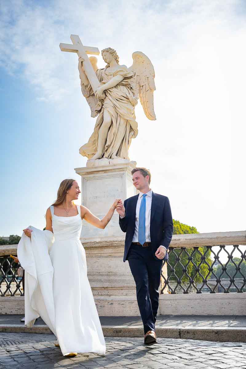Couple walking hand in hand beside a monumental angel sculpture on Ponte Sant’Angelo, captured in a natural walking image along the historic bridge