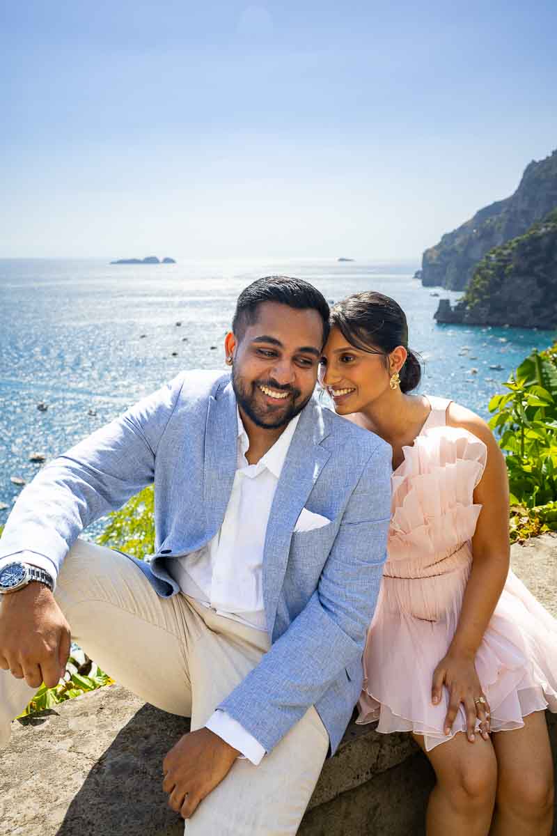 Romantic portrait of a couple sitting on a cliffside wall overlooking the azure Amalfi Coast sea.