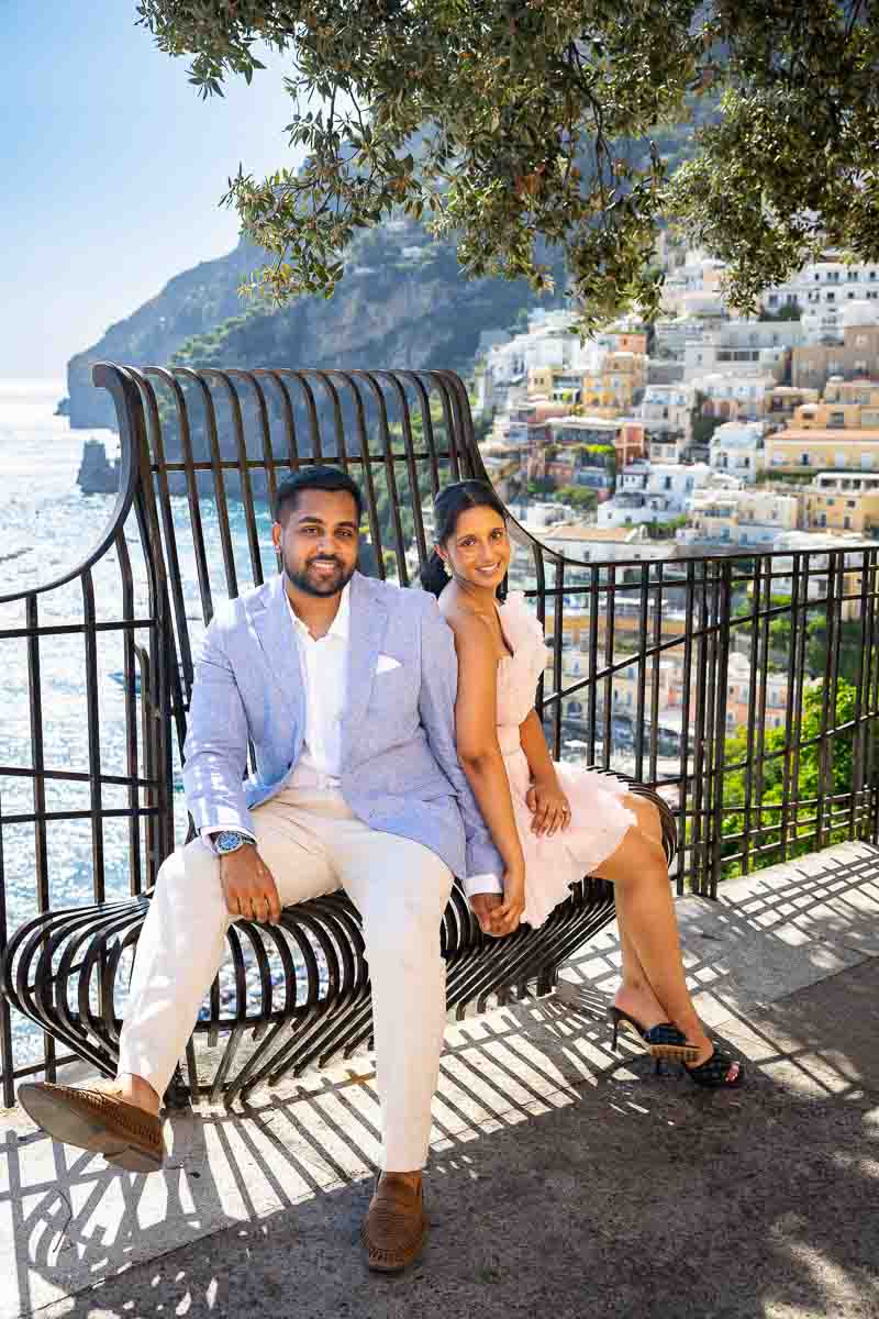 Posed portrait of two lovers sitting on a decorative iron bench with panoramic vistas behind them.